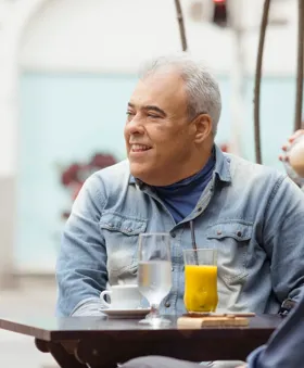 Carlos disfruta de una bebida en la terraza de un café, sonriendo a alguien que no aparece en la imagen.